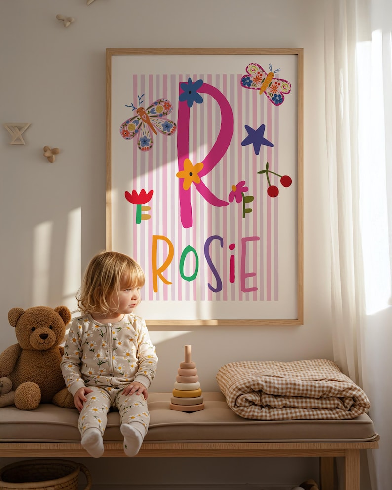 May include: A framed print with the name "Rosie" in colorful letters, decorated with butterflies, flowers, and stars. A young child in a floral onesie sits on a bench with a teddy bear, a wooden stacking toy, and a folded blanket.