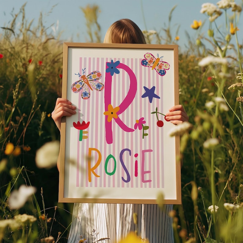 May include: A framed print featuring the name "Rosie" in colorful letters, with a large pink "R" decorated with flowers, butterflies, and a star. The background has pink and white stripes. The print is held in a wooden frame.
