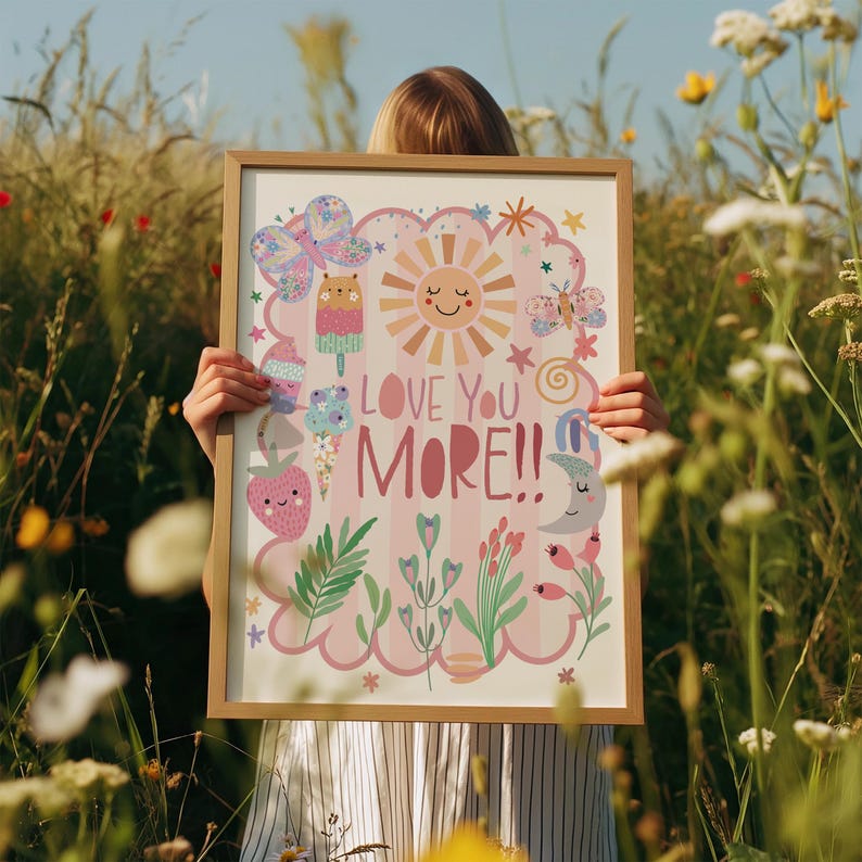 Może przedstawiać: Oprawiony wydruk artystyczny z jasną drewnianą ramą. Wydruk przedstawia r&oacute;żowe tło ze słowami "LOVE YOU MORE!!" oraz ilustracje słońca, motyli, lod&oacute;w, truskawki i element&oacute;w kwiatowych.