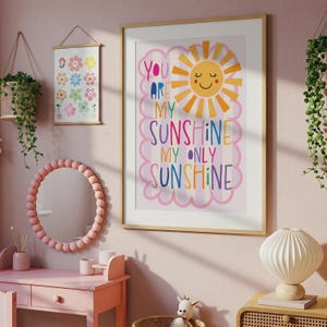 May include: A framed print with the words "You Are My Sunshine, My Only Sunshine" and a smiling sun illustration. The room also features a pink vanity, a round mirror with a beaded frame, and a floral print.