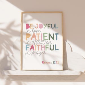 May include: A framed print with a white background and colorful text that reads "Be Joyful in hope, Patient in affliction, Faithful in prayer." The text is in a variety of colors, including pink, orange, yellow, green, blue, and purple. The bottom of the print reads "Romans 12:12".