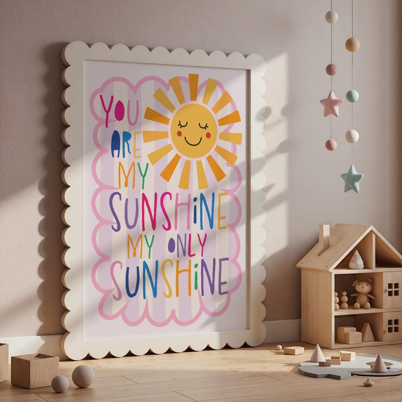 May include: A framed print with the words "You Are My Sunshine My Only Sunshine" in colorful letters. The artwork features a smiling sun and is set against a pink and white striped background. The frame is off-white with a scalloped edge.