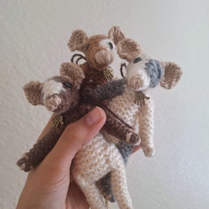 May include: Three crocheted stuffed animals in the shape of pigs. The pigs are white, brown, and gray with black eyes and noses. Each pig has a small gold tag that says "Hand Made".