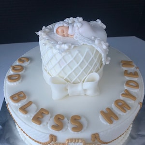 May include: A white cake decorated for a christening. The cake features a sleeping baby figurine in a basket, a bow, and the words "GOD BLESS HAYDEN" in gold lettering. A cross is also featured.