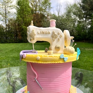 May include: A white and gold sewing machine cake with the name "Linda" on the side. The cake is decorated with pink and yellow frosting and has a spool of thread, needles, and other sewing supplies on top.