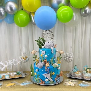 May include: A two-tiered blue birthday cake decorated with sea creatures, surrounded by cupcakes, cookies, and cake pops. Balloons in shades of blue, green, and silver are suspended above the table. A sign on the cake reads "Happy Birthday".