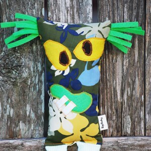 May include: A whimsical fabric doll with a floral pattern in green, yellow, blue, and white. The doll features large yellow eyes with black centers, green felt hair, and striped white legs. A small tag reads "SigiBaby".