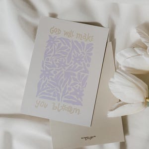 Könnte beinhalten: Eine Grußkarte mit dem Text "God will make you bloom" in goldfarbenen Buchstaben. Die Karte zeigt ein lavendelfarbenes Blumendesign und wird von einem passenden Umschlag begleitet. Weiße Tulpen sind im Hintergrund.