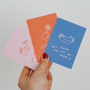 Könnte beinhalten: Drei rechteckige Grußkarten in Hellrosa, Orange und Blau. Die rosa Karte zeigt Hände, die ein Herz formen. Die orangefarbene Karte zeigt eine Hand, die eine Blume hält, und den Text CELEBRATE. Die blaue Karte zeigt eine Umarmung und den Text WIE SCHÖN, DASS WIR UNS HABEN.