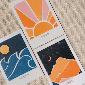 Könnte beinhalten: Drei rechteckige Kunstdrucke mit grafischen Illustrationen. Einer zeigt einen orangefarbenen Sonnenaufgang, ein anderer blaue Meereswellen mit einer gelben Sonne und der dritte orangefarbene Berge unter einer gelben Mondsichel. Jeder Druck enthält deutschen Text.