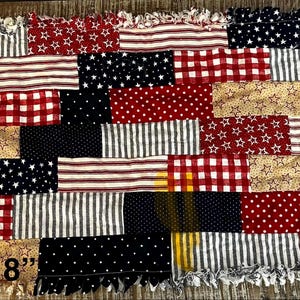 May include: A patchwork rectangular mat, 11" x 18", with a variety of patriotic patterns. The design includes red and white gingham, navy blue with white stars, and tan with white stars. The edges have a fringed trim.