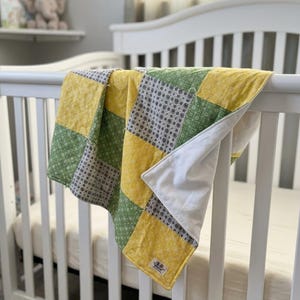 May include: A patchwork baby quilt draped over a white crib. The quilt features squares of yellow, green, and patterned gray fabric. The quilt is made of soft material and has a white backing.