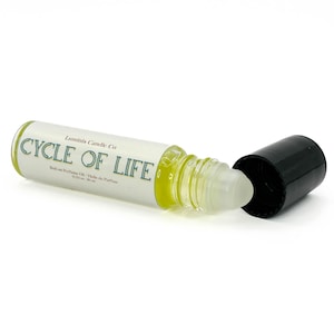 May include: A roll-on perfume oil bottle with the text "CYCLE OF LIFE" in green lettering. The bottle is mostly white with a green border and contains a light yellow liquid. The black cap is off to the side.