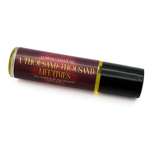 May include: A roll-on perfume oil bottle with a dark burgundy label featuring gold lettering that reads "A Thousand Thousand Lifetimes." The bottle has a black cap and a clear glass body, with a yellow liquid visible inside. The product is from Luminis Candle Co.