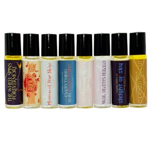 May include: A collection of eight roll-on perfume oils with black caps and colorful labels. Each label features a unique name and design, including text such as "The Wheel Spins Forevermore" and "Everything is in Everything."
