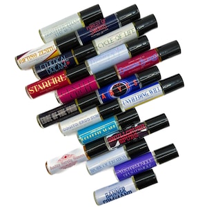 May include: An assortment of roll-on perfume bottles with black lids and labels. The labels display phrases such as "Brighter Than The Sun," "Forest of Swords," and "Cycle of Life." The bottles are arranged on a white surface.