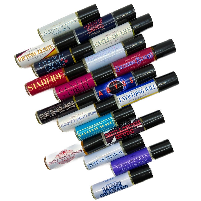 May include: An assortment of roll-on perfume bottles with black lids and labels. The labels display phrases such as "Brighter Than The Sun," "Forest of Swords," and "Cycle of Life." The bottles are arranged on a white surface.