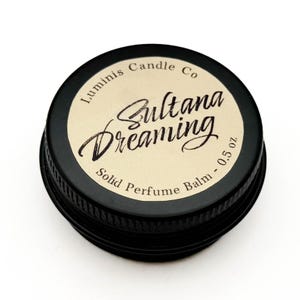 Puede incluir: Un recipiente redondo negro de bálsamo de perfume sólido. La etiqueta dice "Luminis Candle Co" y "Sultana Dreaming". La etiqueta es de color canela con texto negro y el recipiente es de 14 gramos.