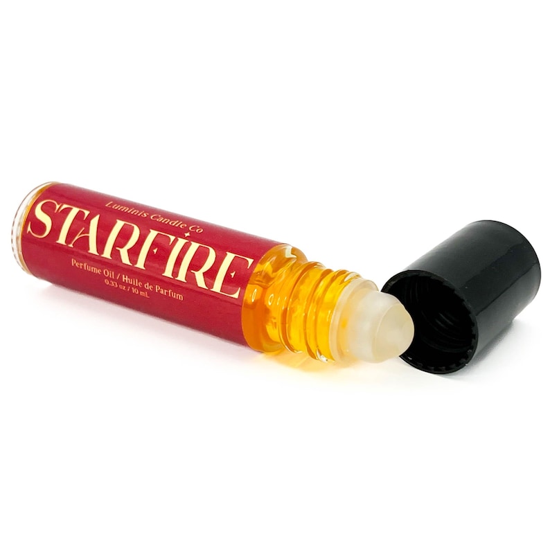 May include: A red and gold perfume oil roller bottle with the word "STARFIRE" in gold lettering. The bottle is open, revealing a clear rollerball applicator and a black cap. The label also reads "Perfume Oil / Huile de Parfum 0.33 fl oz / 10 ml."