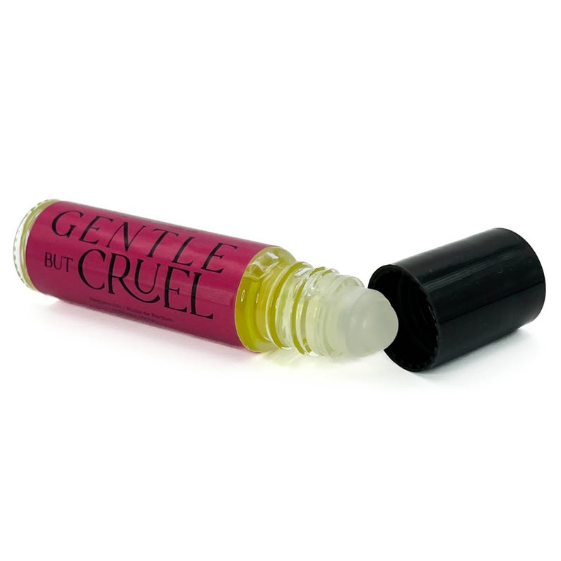 May include: A clear glass roll-on bottle with a pink label reading "GENTLE BUT CRUEL" in white text. The bottle contains a yellow liquid and has a black cap lying next to it. The roll-on applicator is visible.