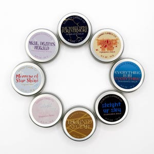 May include: A collection of ten small, round fragrance balm tins arranged in a circle. Each tin has a silver lid and a label with text such as "The Wheel Spins Forevermore" and "Everything is in Everything". Each tin is 0.5 oz / 14 g.
