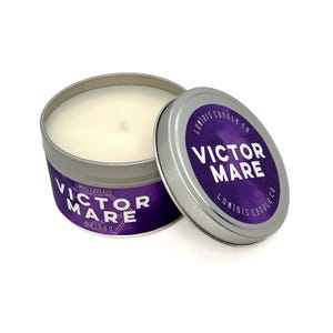 Puede incluir: Una vela en una lata de plata con una etiqueta morada que dice "Luminis Candle Co. Victor Mare Beidou". La vela está en una lata de plata con una tapa que tiene la misma etiqueta.