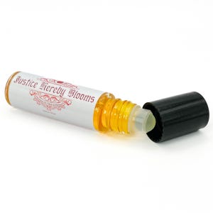 May include: A clear glass roll-on bottle with a black cap, containing a golden-yellow liquid. The bottle has a white label with the text "Justice Hereby Blooms". The cap is off, revealing the rollerball applicator. The bottle is designed for perfume.
