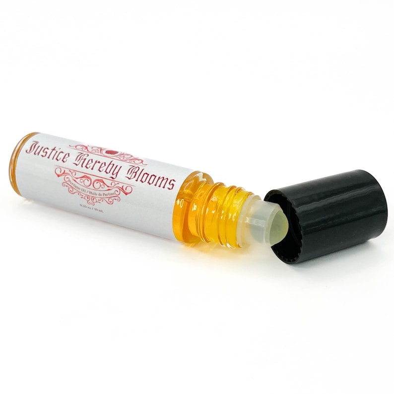 May include: A clear glass roll-on bottle with a black cap, containing a golden-yellow liquid. The bottle has a white label with the text "Justice Hereby Blooms". The cap is off, revealing the rollerball applicator. The bottle is designed for perfume.