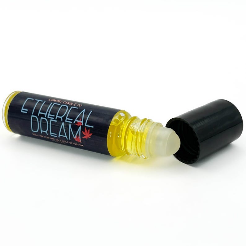 May include: A roll-on perfume bottle with a black label displaying "Ethereal Dream" in white. The bottle contains a yellow liquid and has a black cap. The label also features red leaf designs.