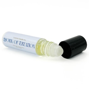 May include: A clear glass roll-on bottle with a white label that reads "WORK OF CREATION" in blue text. The black cap is off, revealing the rollerball applicator. The bottle contains a light yellow liquid.