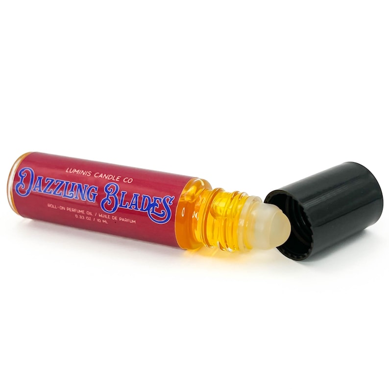 May include: A roll-on perfume oil bottle with a red label and the text "DAZZLING BLADES" in blue. The bottle contains a yellow liquid and has a black cap. The label also includes "LUMINIS CANDLE CO."