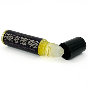 May include: A roll-on perfume oil bottle with the text "Edge of the Void" in white lettering on a black label. The bottle contains a yellow liquid and has a clear rollerball applicator. The black cap is off to the side.