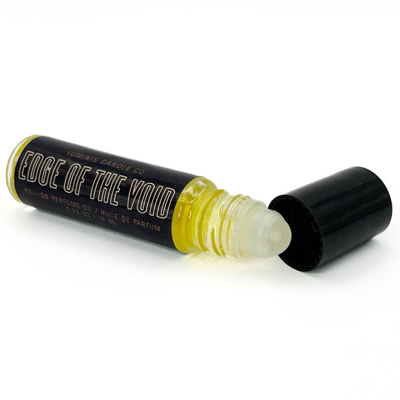 May include: A roll-on perfume oil bottle with the text "Edge of the Void" in white lettering on a black label. The bottle contains a yellow liquid and has a clear rollerball applicator. The black cap is off to the side.