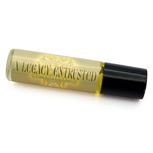 May include: A roll-on perfume bottle with a black cap and a clear glass body. The bottle has a gold label with the text "A Legacy Entrusted" and "Luminis Candle Co." The liquid inside is yellow.
