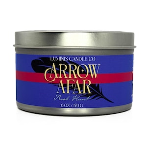 Puede incluir: Una lata de vela con tapa plateada y una etiqueta azul con el texto "ARROW AFAR" en letras doradas, un gráfico de pluma negra y una franja roja. La etiqueta también incluye "LUMINIS CANDLE CO" y "6 OZ / 170 G".