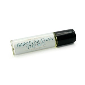 May include: A clear glass roll-on bottle with a black cap. The label reads "BRIGHTER THAN THE SUN" in teal lettering. The bottle contains a light yellow liquid, likely perfume or essential oil. The product is from Luminis Candle Co.