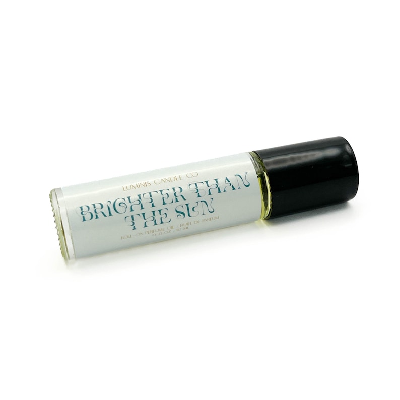 May include: A clear glass roll-on bottle with a black cap. The label reads "BRIGHTER THAN THE SUN" in teal lettering. The bottle contains a light yellow liquid, likely perfume or essential oil. The product is from Luminis Candle Co.