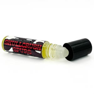 May include: A roll-on perfume bottle with the text "SKULLCRUSH SPURS" in red and white lettering. The bottle has a black cap and a clear rollerball applicator. The label has a black and white cowhide pattern.