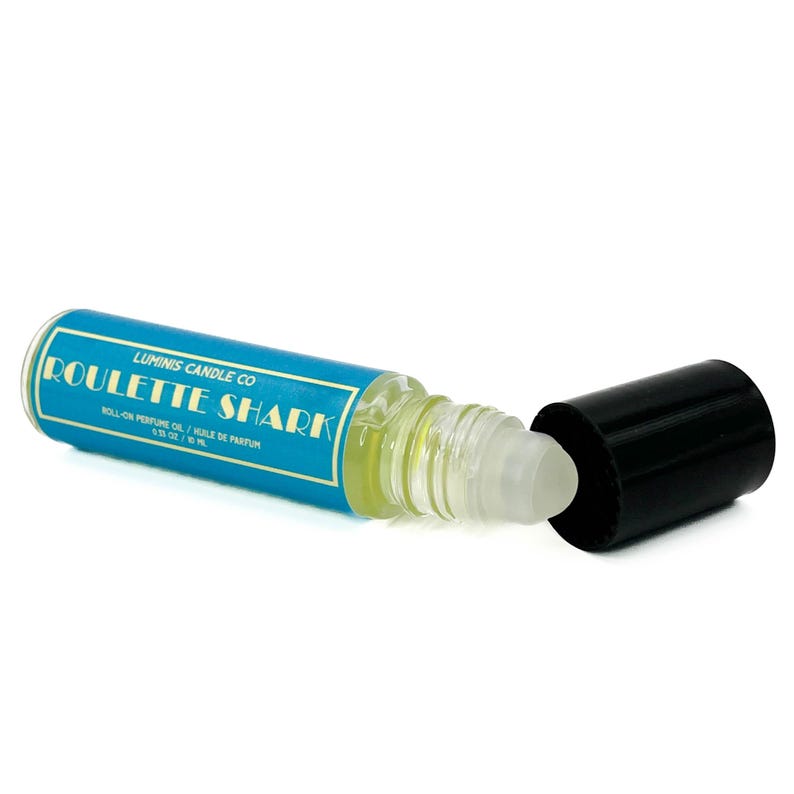May include: A roll-on perfume oil bottle with a blue label reading "Roulette Shark" and "Luminis Candle Co." The clear bottle contains yellow liquid. The black cap is off to the side. The bottle is 10 ml.