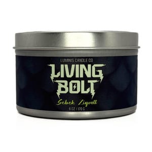 May include: A silver metal candle tin with a black label featuring the text "LIVING BOLT" in a stylized font with a lightning bolt graphic. The label also includes "Sebek Zigvolt" and "6 OZ / 170 G". The candle is from Luminis Candle Co.