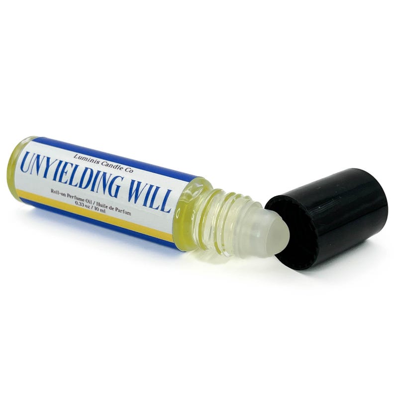 May include: A clear roll-on perfume oil bottle with the text "UNYIELDING WILL" in blue and white. The bottle contains a yellow liquid. The black cap is off, showing the rollerball. The product is from Luminis Candle Co.