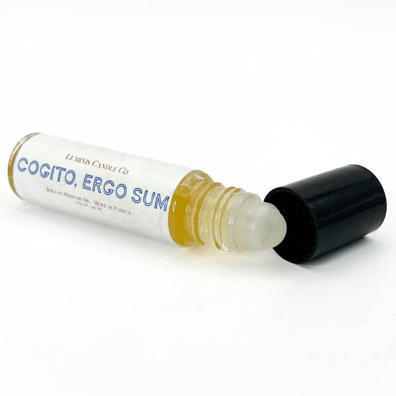May include: A clear glass roll-on perfume oil bottle with a black cap. The bottle contains a yellow liquid and has a white label with blue text that reads "COGITO, ERGO SUM" and "LUMINUS CANDLE CO."