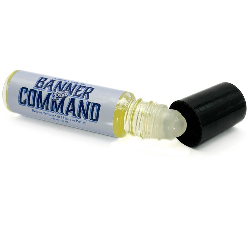 May include: A clear glass roll-on perfume oil bottle with a black cap. The bottle has a white label with the words "BANNER OF COMMAND" in blue. The bottle contains a yellow liquid. The cap is off, revealing the rollerball applicator.