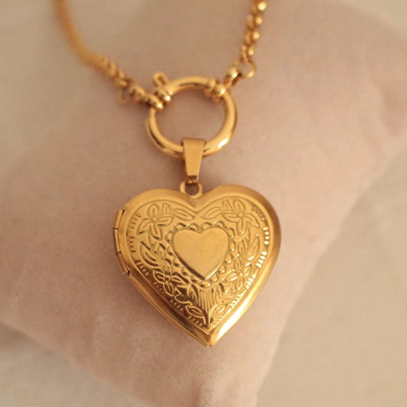 May include: A gold heart-shaped locket necklace with a delicate chain and a floral design. The locket has a small heart-shaped opening in the center.