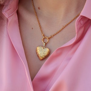May include: A gold chain necklace with a heart-shaped locket pendant. The locket has an intricate floral design.