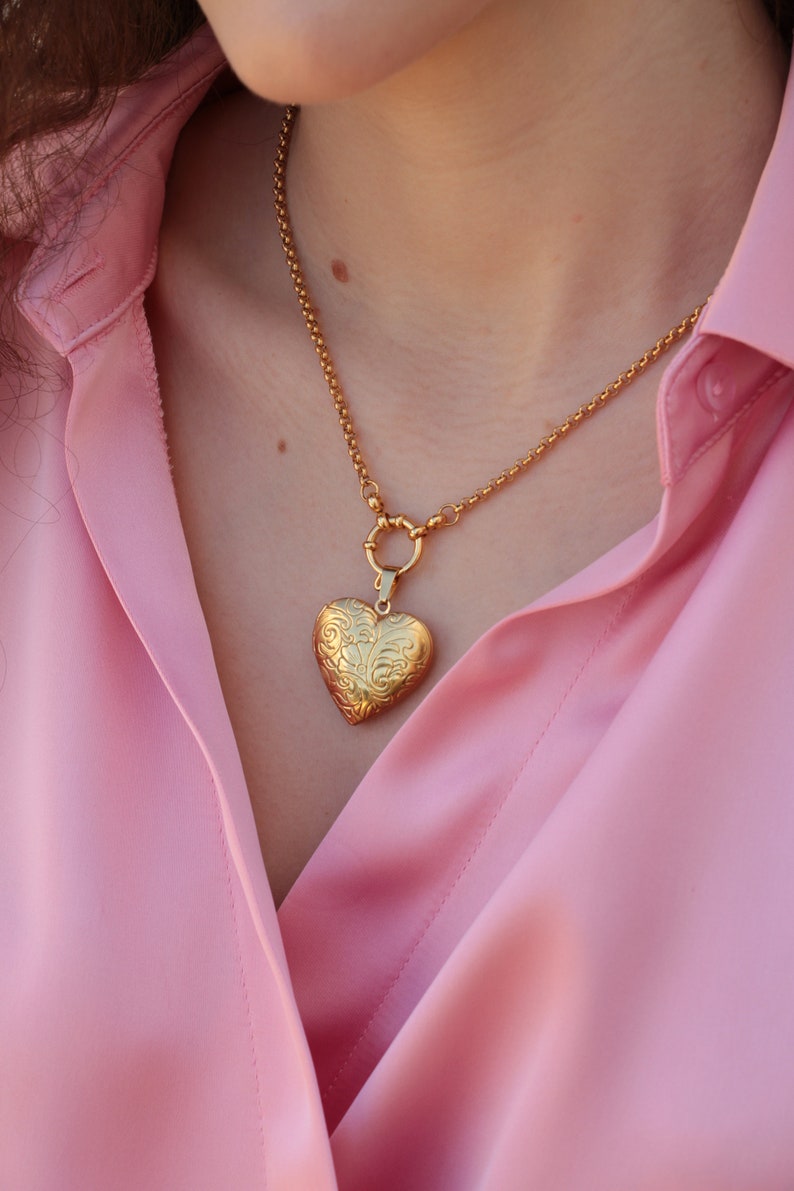 May include: A gold chain necklace with a heart-shaped locket pendant. The locket has an intricate floral design.