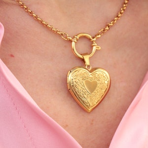 May include: A gold chain necklace with a heart-shaped locket pendant. The locket has a detailed floral design and a smaller heart in the center.