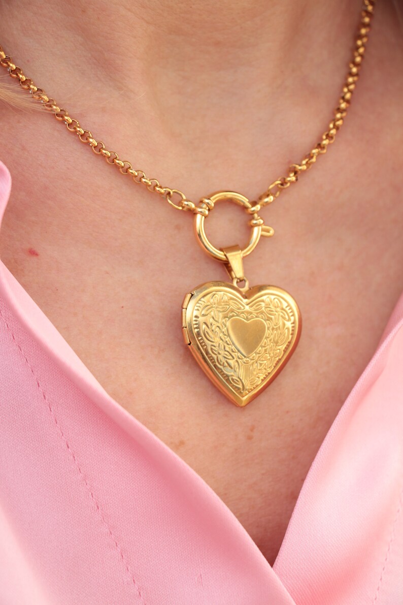 May include: A gold chain necklace with a heart-shaped locket pendant. The locket has a detailed floral design and a smaller heart in the center.