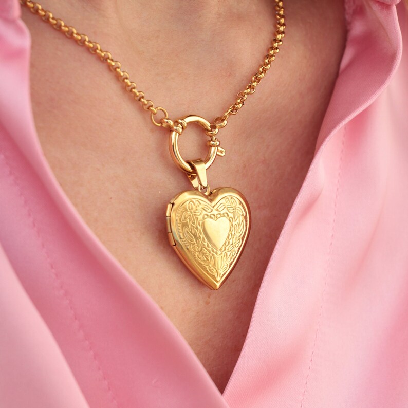 May include: A gold chain necklace with a heart-shaped locket pendant. The locket has an intricate design and a smaller heart in the center.