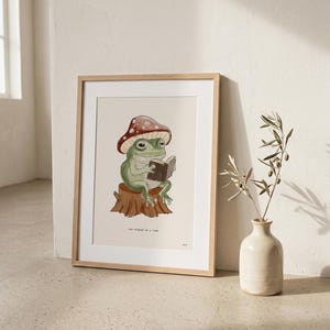 May include: Framed art print featuring a whimsical illustration of a green frog wearing a mushroom cap and reading a book while seated on a tree stump. The text "One chapter at a time" is below the image. The frame is light wood.